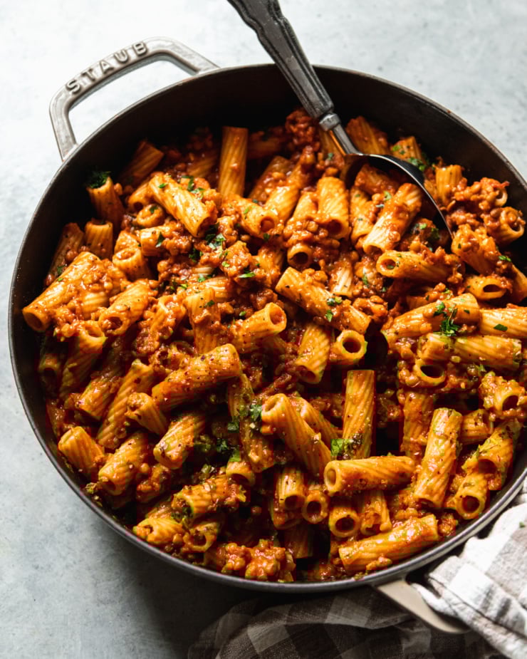 An overhead shot of a braiser pot filled with rigatoni with lentil bolognese sauce. The pasta is garnished with finely chopped parsley and a spoon is sticking out of the pot.
