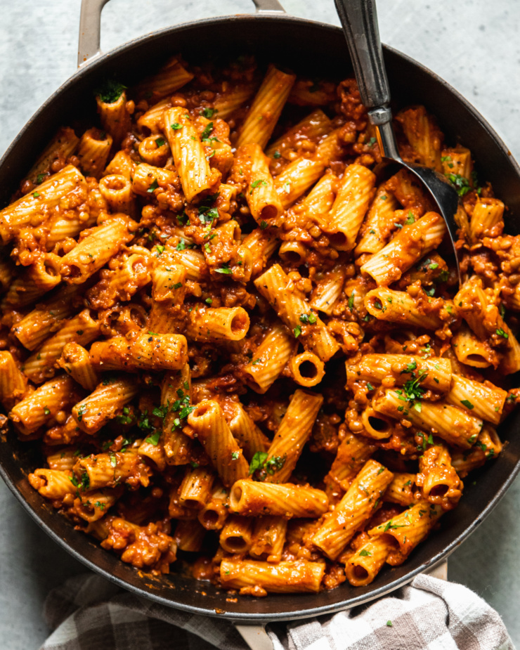 An up close, overhead shot of a braiser pot filled with rigatoni with lentil bolognese sauce. The pasta is garnished with finely chopped parsley and a spoon is sticking out of the pot.