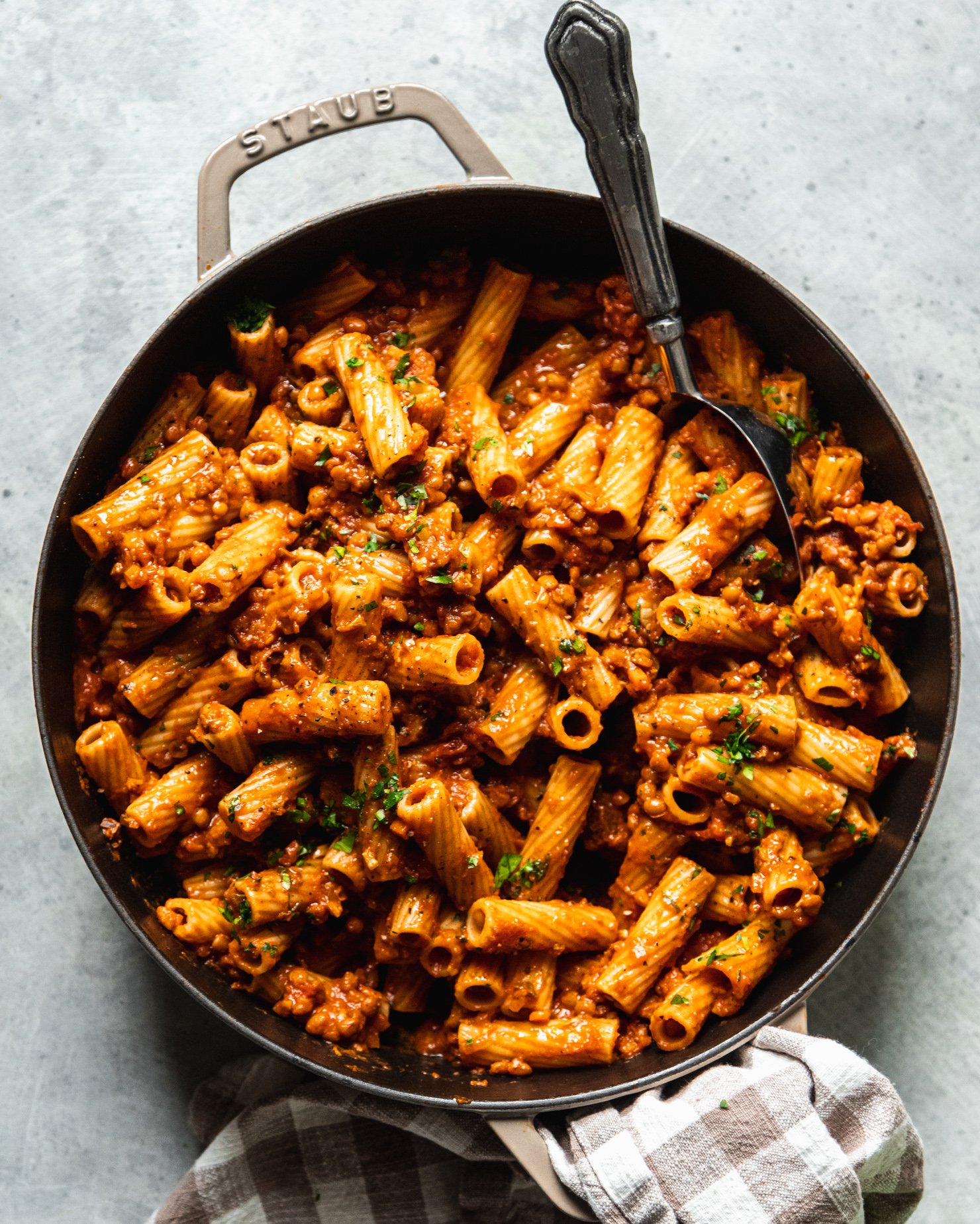 An overhead shot of a braiser pot filled with rigatoni with lentil bolognese sauce. The pasta is garnished with finely chopped parsley and a spoon is sticking out of the pot.