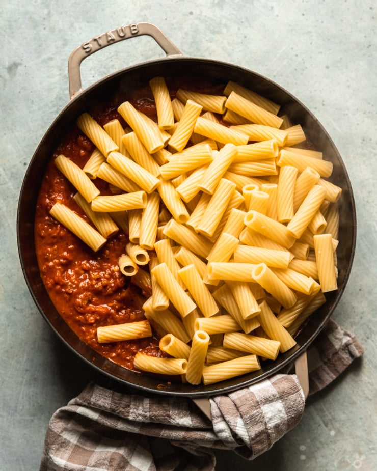 An overhead shot shows freshly cooked rigatoni piled on top of a lentil bolognese sauce in a braiser-style pot. A napkin is wrapped around the pot's handle.