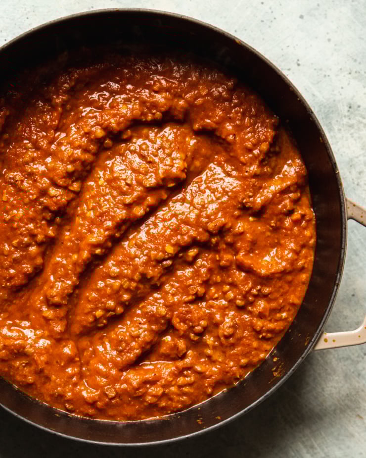 An up close, overhead shot shows a braiser-style pot filled with a lentil bolognese sauce. The pot handle is splattered with tomato concentrate.