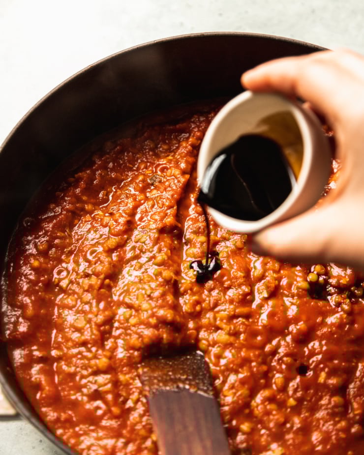 A slight 3/4 angle shot shows a hand pouring a small bowl of balsamic vinegar into a wide pot filled with a tomato-y sauce.