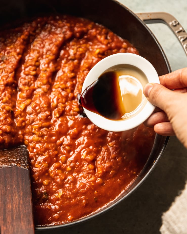 An overhead shot shows a hand emptying a small bowl of Tamari into a wide pot filled with a tomato-y lentil sauce.
