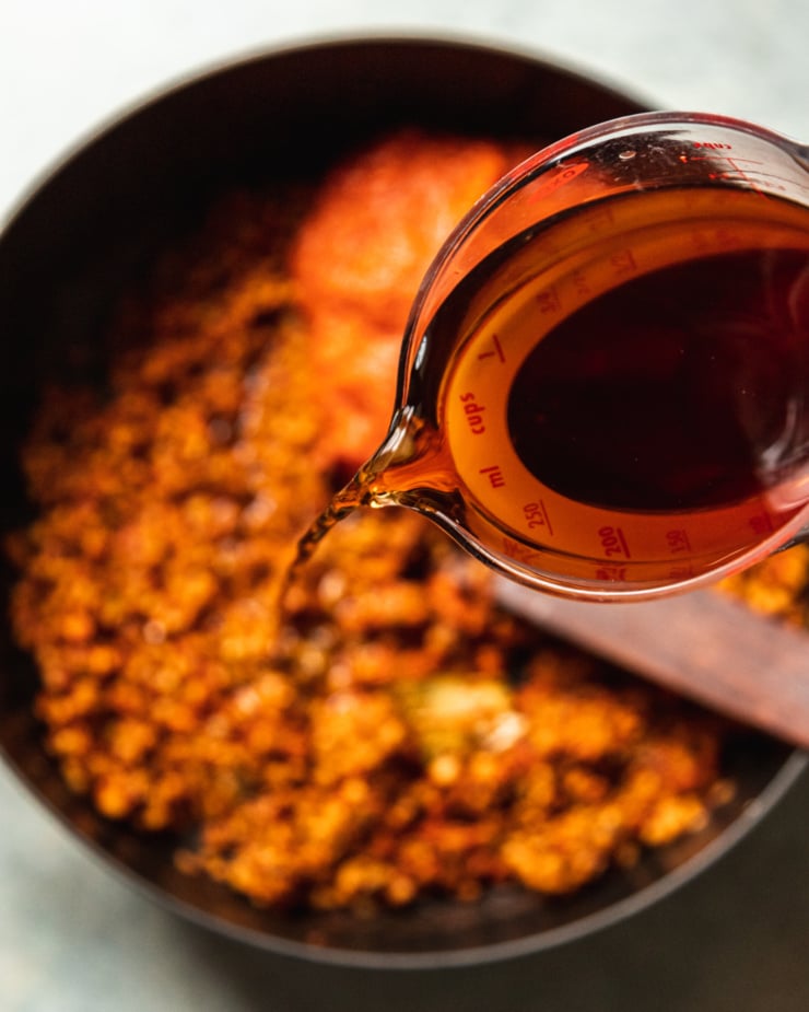 An overhead shot shows a measuring cup emptying dried porcini soaking liquid into a large pot of lentil bolognese sauce.
