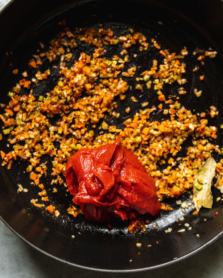 An overhead shot shows sautéed carrots, celery and shallots in a pot with a dollop of tomato paste that has just been added.