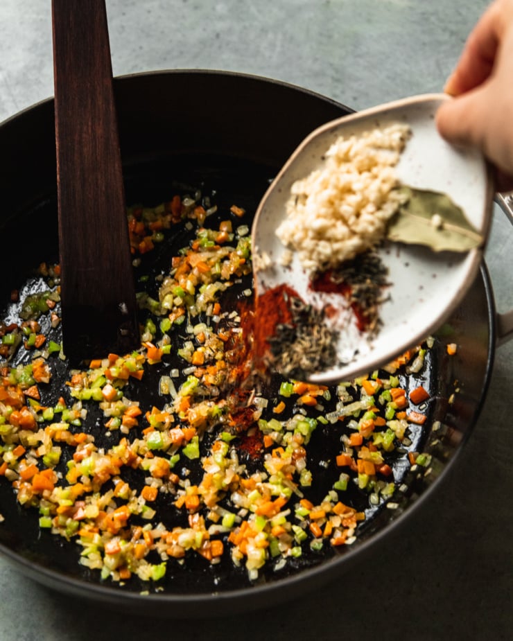 A slight 3/4 angle shot shows garlic, smoked paprika, thyme, and a bay leaf being added to a pot with sautéed celery, carrots, and shallots.