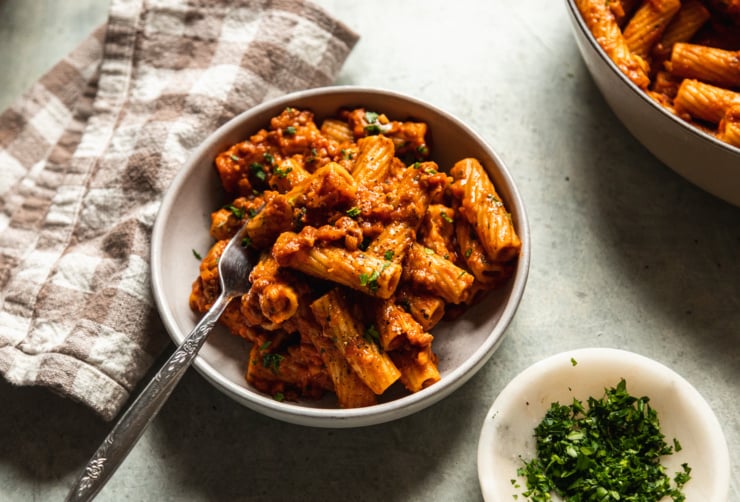 A 3/4 angle shot of a serving of lentil bolognese rigatoni in a wide, white bowl. A checkered napkin is nearby and the pasta is garnished with finely chopped parsley.