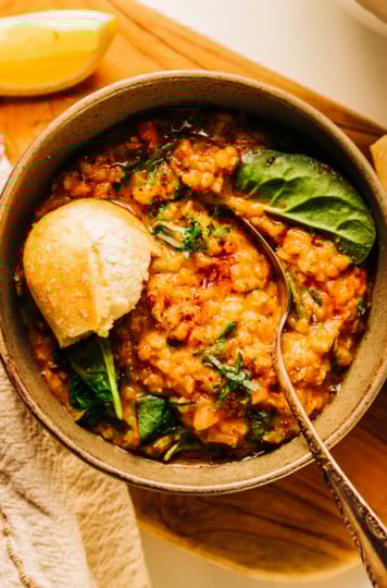 An overhead shot of a bowl of lemony lentil soup garnished with chopped parsley and a crust of baguette sticking out. There is a lemon slice and linen napkin nearby.
