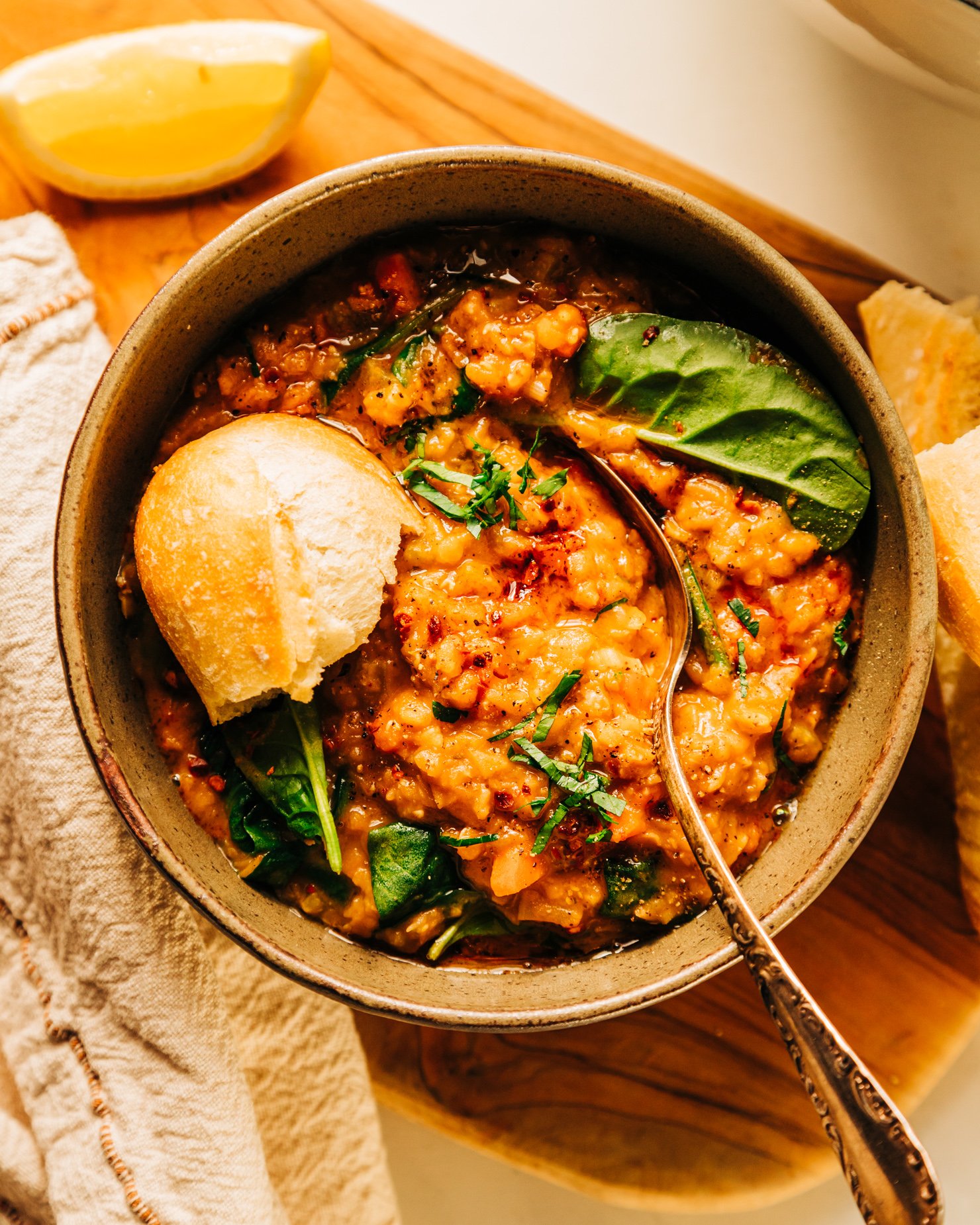An overhead shot of a bowl of lemony lentil soup garnished with chopped parsley and a crust of baguette sticking out. There is a lemon slice and linen napkin nearby.