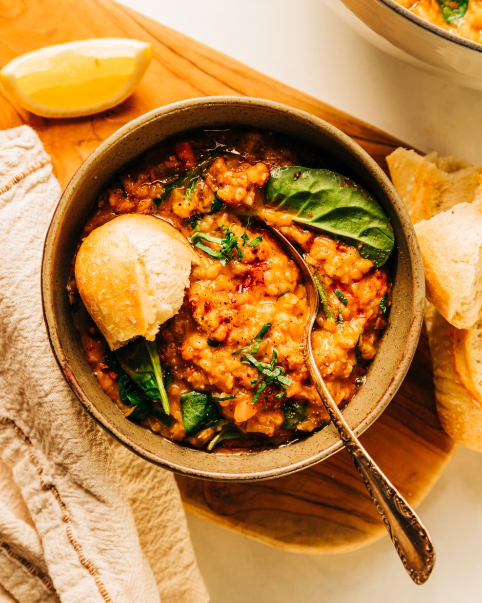 An overhead shot of a bowl of lemony lentil soup garnished with chopped parsley and a crust of baguette sticking out. There is a lemon slice and linen napkin nearby.