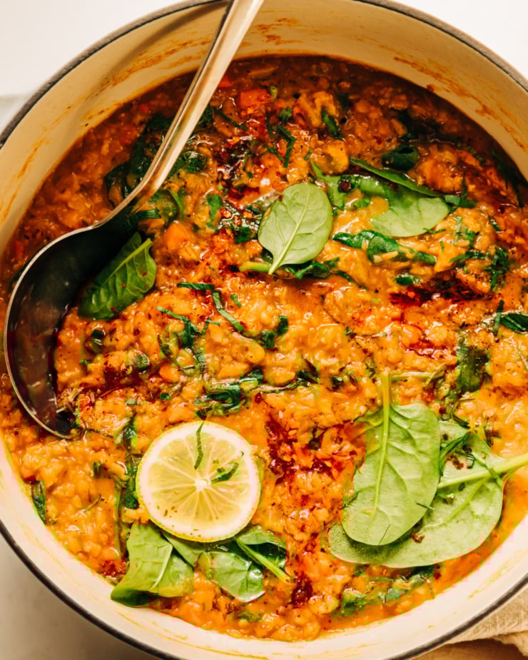 An overhead shot of a pot of lemony lentil soup with spinach. The soup is garnished with chopped parsley and chili crisp. A ladle is sticking out of the pot of soup.