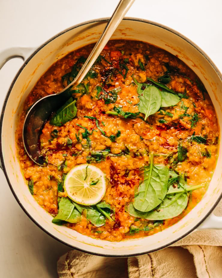 An overhead shot of a pot of lemony lentil soup with spinach. The soup is garnished with chopped parsley and chili crisp. A ladle is sticking out of the pot of soup.