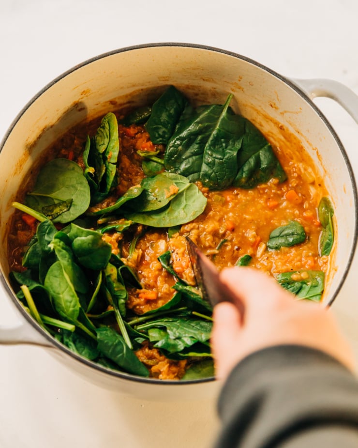An overhead shot of a hand using a wooden utensil to stir some spinach into a pot of lentil soup.