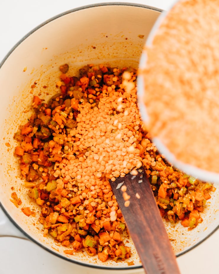An overhead shot shows red split lentils being added to a pot of sautéed and spiced vegetables.