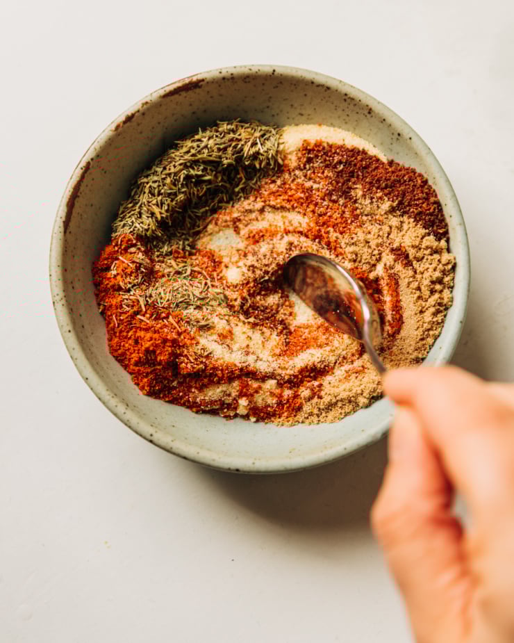 An overhead shot shows a hand using a spoon to stir together a blend of 7 spices.