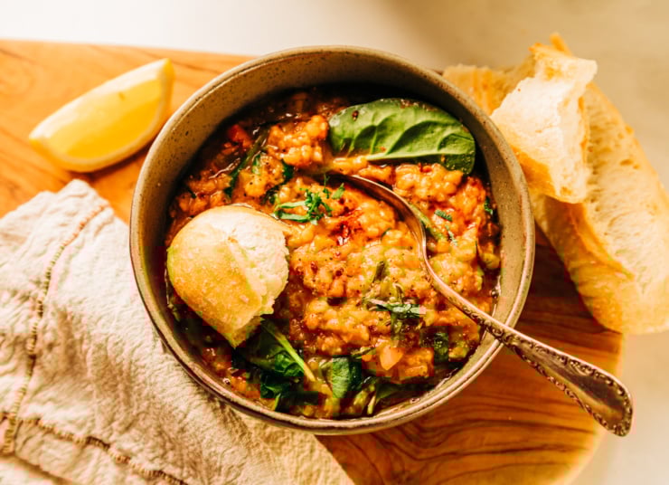 An overhead shot of a bowl of lemony lentil soup garnished with chopped parsley and a crust of baguette sticking out. There is a lemon slice and linen napkin nearby.
