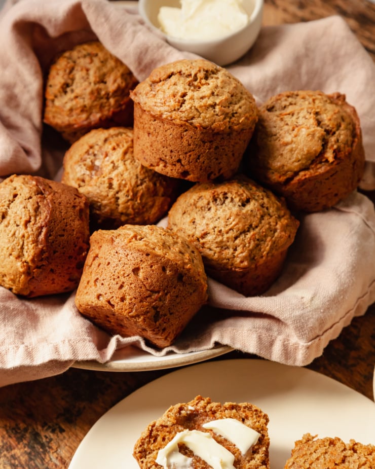 A slight 3/4 angle shot shows vegan carrot muffins in a linen-lined bowl. The muffins are golden brown with flecks of carrot, and have nicely domed tops.