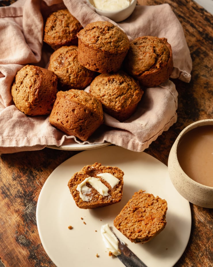 A slight 3/4 angle shot shows vegan carrot muffins in a linen-lined bowl. The muffins are golden brown with flecks of carrot, and have nicely domed tops.