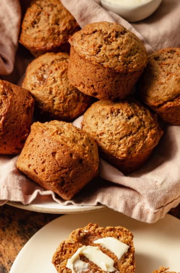 An up close, slight 3/4 angle shot shows vegan carrot muffins in a linen-lined bowl. The muffins are golden brown with flecks of carrot, and have nicely domed tops.