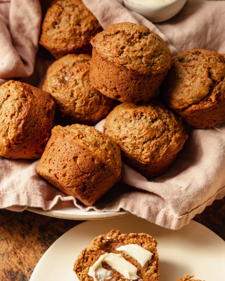 An up close, slight 3/4 angle shot shows vegan carrot muffins in a linen-lined bowl. The muffins are golden brown with flecks of carrot, and have nicely domed tops.