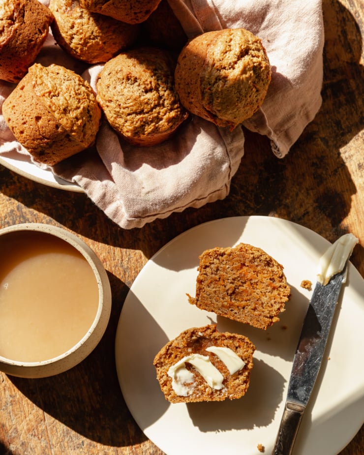 An overhead shot shows vegan carrot muffins in a linen-lined bowl. The muffins are golden brown with flecks of carrot, and have nicely domed tops. A split muffin is on a plate to the side where half of it is swopped with some vegan butter. There is also a cup of tea nearby.
