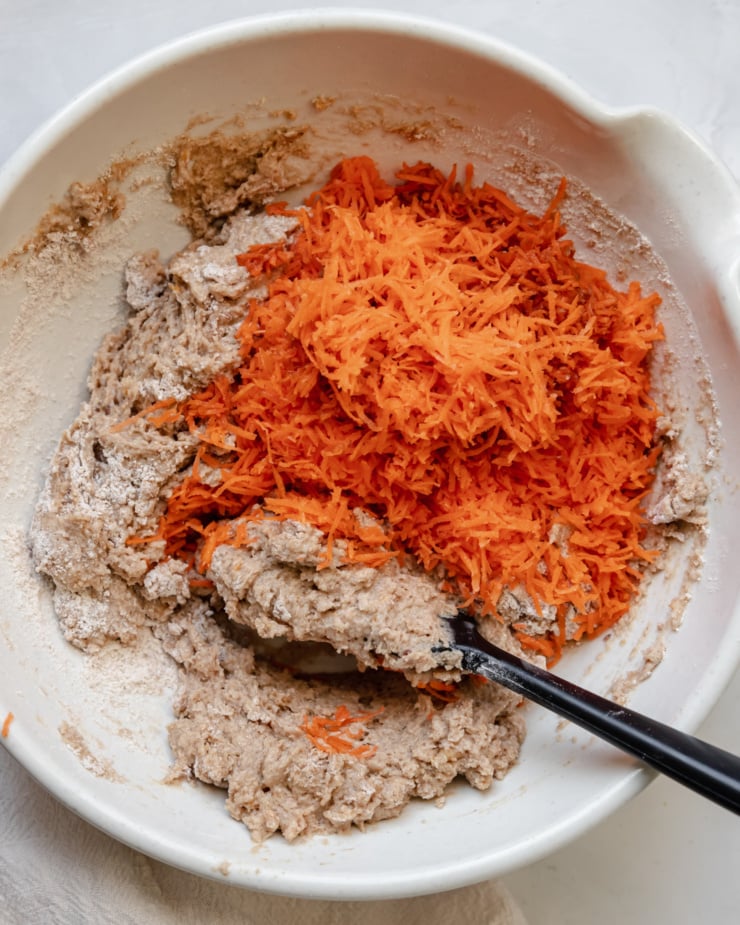 An overhead shot shows shredded carrots being added to a bowl of muffin batter.