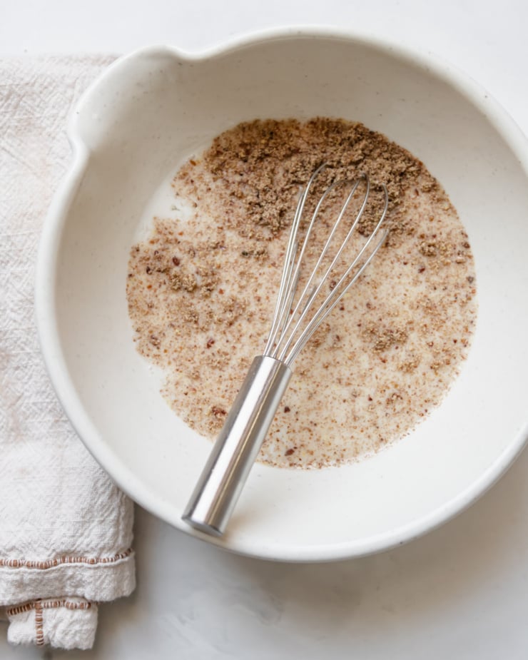 An overhead shot of a small mixing bowl with some ground flax seeds sprinkled on top. A small whisk is sticking out of the liquid in the bowl.