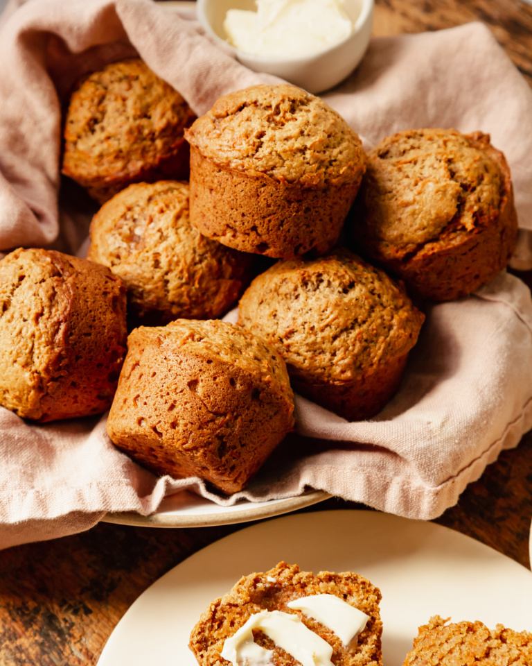 A slight 3/4 angle shot shows vegan carrot muffins in a linen-lined bowl. The muffins are golden brown with flecks of carrot, and have nicely domed tops.