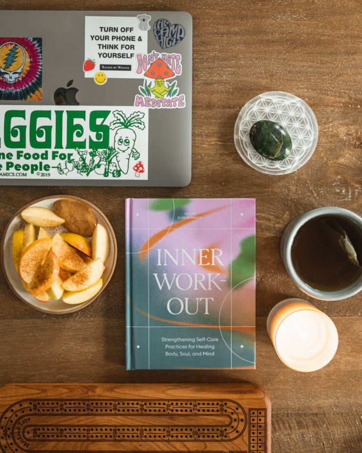 An overhwad shot shows a laptop computer adorned with stickers, a selenite coaster, a polished piece of jade, a small plate with a sliced apple on it, a copy of the book "Inner Workout" by Taylor Elyse Morrison, a cup of tea, a lit candle, and a wooden cribbage board.