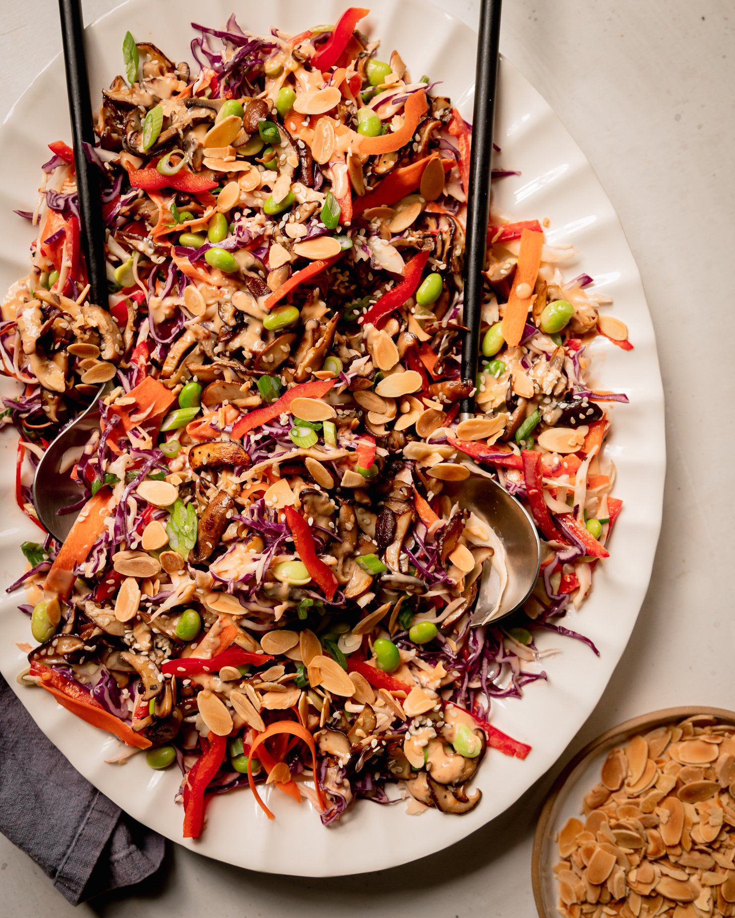 An overhead shot of a platter of spicy sesame slaw topped with sautéed mushrooms and shelled edamame. The slaw is garnished with toasted sliced almonds. A set of salad tongs is sticking out of the slaw and a linen napkin is nearby.