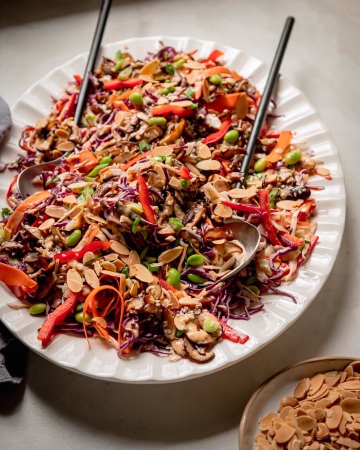 A 3/4 angle shot of a platter of spicy sesame slaw topped with sautéed mushrooms and shelled edamame. The slaw is garnished with toasted sliced almonds. A set of salad tongs is sticking out of the slaw and a linen napkin is nearby.