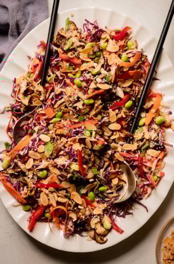 An overhead shot of a platter of spicy sesame slaw topped with sautéed mushrooms and shelled edamame. The slaw is garnished with toasted sliced almonds. A set of salad tongs is sticking out of the slaw and a linen napkin is nearby.