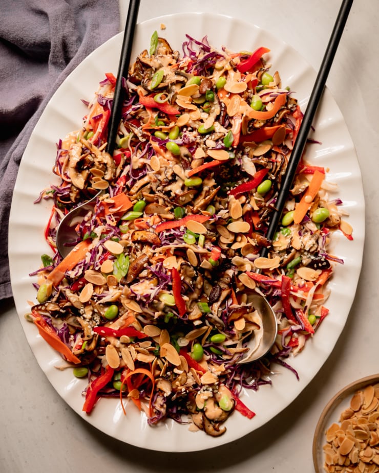 An overhead shot of a platter of spicy sesame slaw topped with sautéed mushrooms and shelled edamame. The slaw is garnished with toasted sliced almonds. A set of salad tongs is sticking out of the slaw and a linen napkin is nearby.
