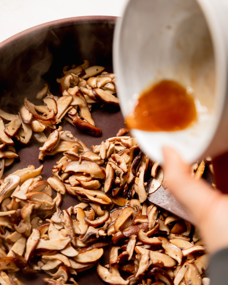 An overhead shot shows a hand pouring a mixture into a steaming skillet of sautéed mushrooms.