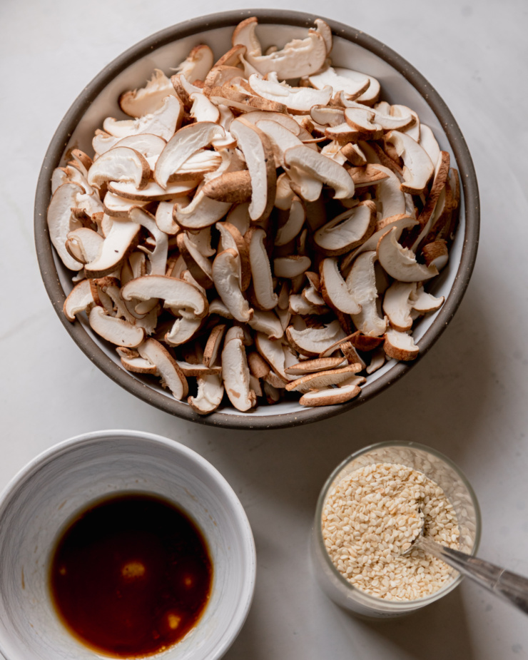 An overhead shot shows a bowl of sliced mushrooms, a small bowl with some maple syrup and Tamari in it, and a small jar of sesame seeds.