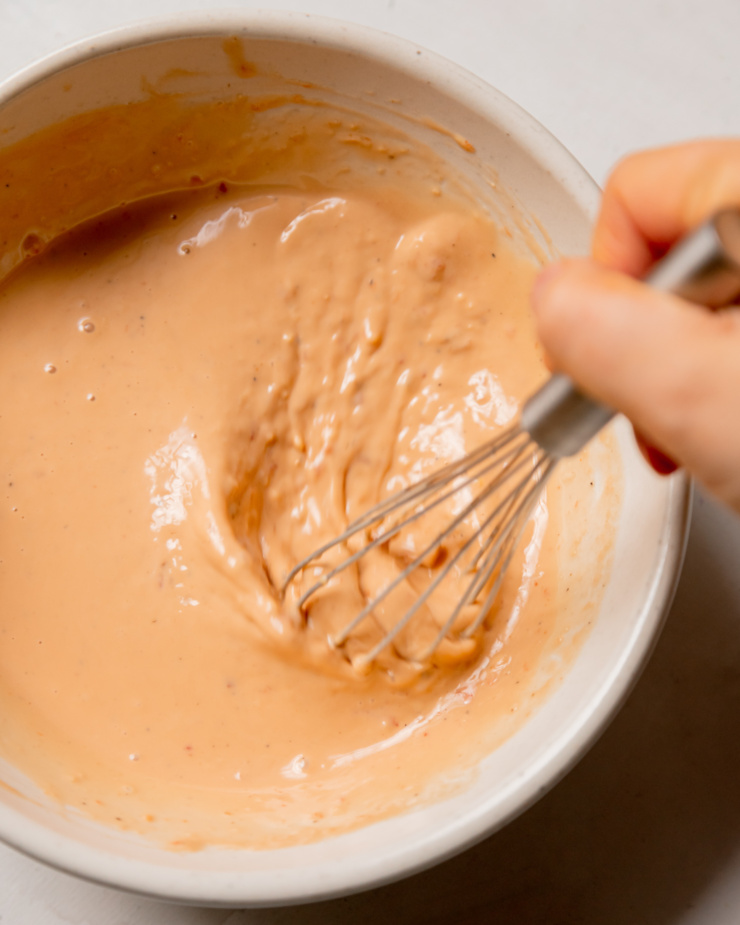An up close, overhead shot shows a hand using a small whisk to stir up a spicy sesame dressing that is creamy in texture.