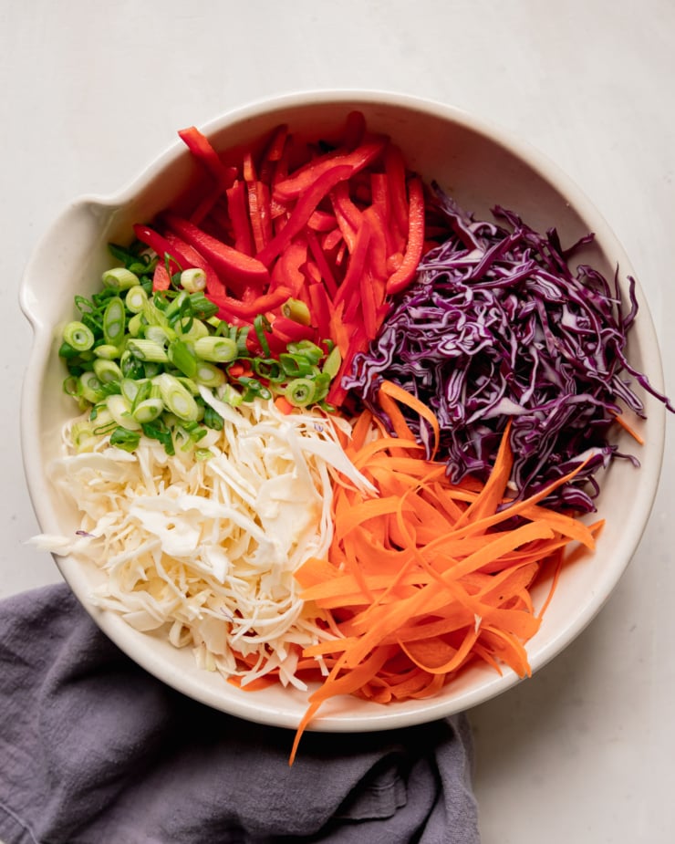 An overhead shot of the shredded/sliced/prepped components of a spicy sesame slaw: red bell pepper, red cabbage, carrot, green cabbage, and green onions.