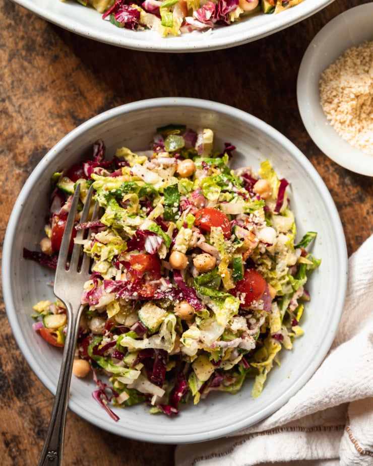 An overhead shot of a singular serving of a pizza night salad in a wide, white and speckled bowl. The salad contains: romaine, radicchio, chickpeas, olives, pepperoncini, sun dried tomatoes, red onion, cucumber, cherry tomatoes, and a red wine vinegar and thyme dressing.