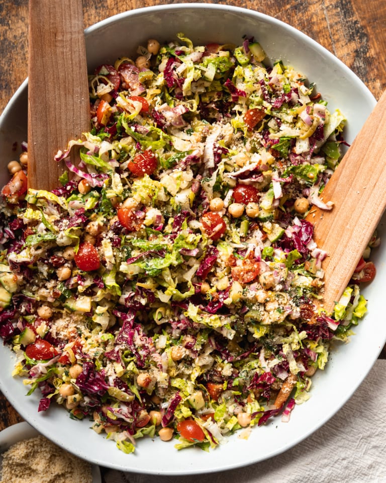 An overhead shot of a pizza night salad in a wide, white bowl with wooden salad tongs. The salad contains: romaine, radicchio, chickpeas, olives, pepperoncini, sun dried tomatoes, red onion, cucumber, cherry tomatoes, and a red wine vinegar and thyme dressing.