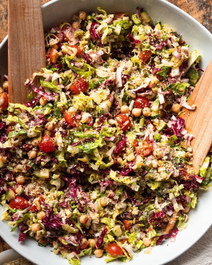 An overhead shot of a pizza night salad in a wide, white bowl with wooden salad tongs. The salad contains: romaine, radicchio, chickpeas, olives, pepperoncini, sun dried tomatoes, red onion, cucumber, cherry tomatoes, and a red wine vinegar and thyme dressing.
