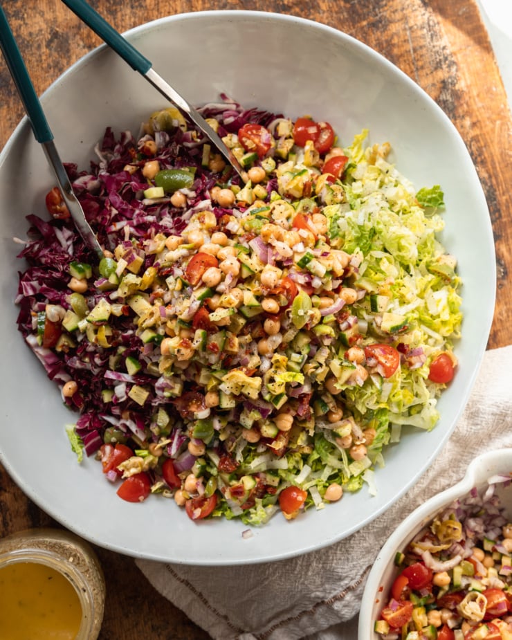 An overhead shot shows a salad being assembled in a wide white bowl over a worn wooden surface. The dressing is in a jar nearby.
