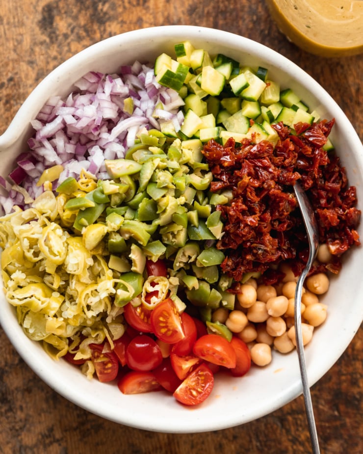An overhead shot of a wide white bowl containing segments of: red onion, cucumber, sun dried tomatoes, chickpeas, cherry tomatoes, pepperoncini peppers, and green olives. Everything is chopped finely.
