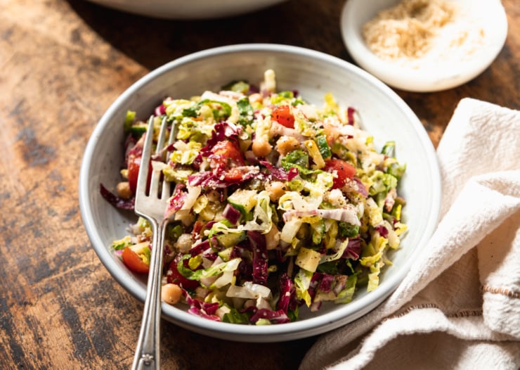A 3/4 angle shot of a singular serving of a pizza night salad in a wide, white and speckled bowl. The salad contains: romaine, radicchio, chickpeas, olives, pepperoncini, sun dried tomatoes, red onion, cucumber, cherry tomatoes, and a red wine vinegar and thyme dressing.