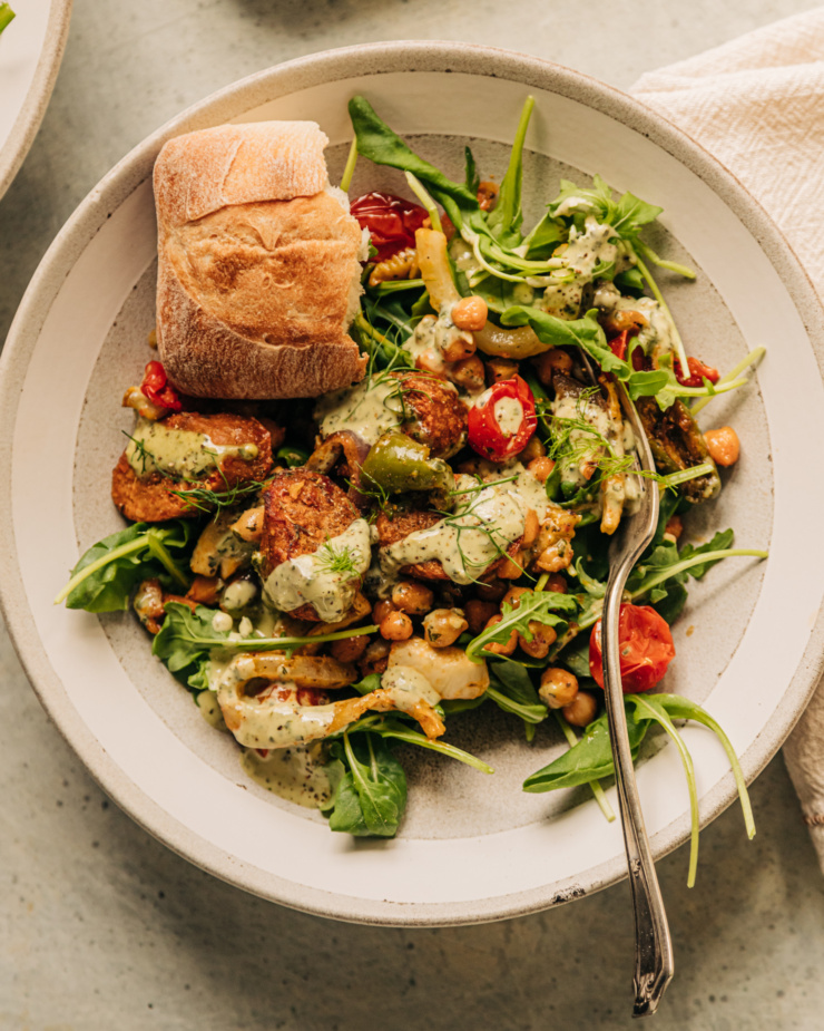 An up close, overhead shot shows a serving of veggie sausage sheet pan dinner served on top of arugula in a serving bowl. A crust of ciabatta bread is on the side in the bowl along with extra drizzles of vegan pesto cream sauce.