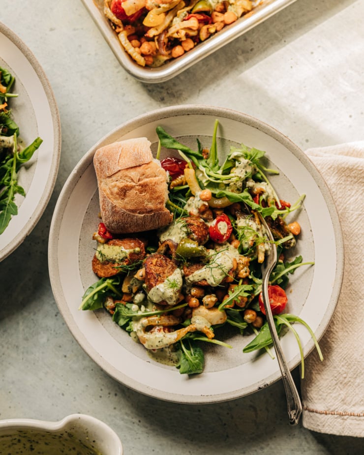 An overhead shot shows a serving of veggie sausage sheet pan dinner served on top of arugula in a serving bowl. A crust of ciabatta bread is on the side in the bowl along with extra drizzles of vegan pesto cream sauce.