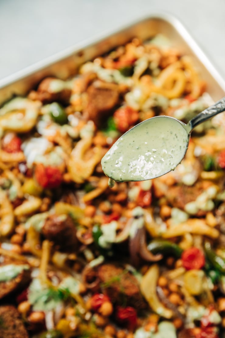 A 3/4 angle image shows a spoon dripping a vegan pesto cream sauce on top of a tray of roasted veggies and vegan sausage.