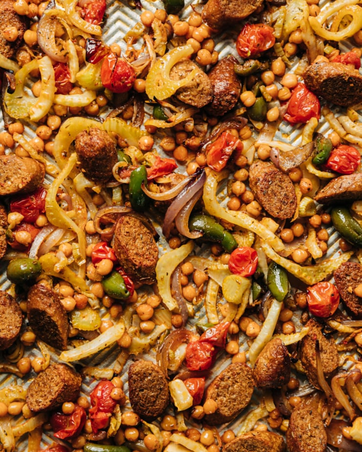 An up close, overhead shot shows roasted onions, fennel, chickpeas, cherry tomatoes, and sliced vegan sausage on a baking sheet.