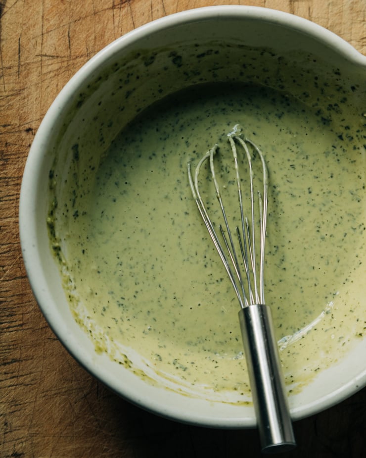 An up close, overhead shot shows a bowl of vegan pesto cream sauce with a whisk. The sauce is light green and flecked with darker green bits.