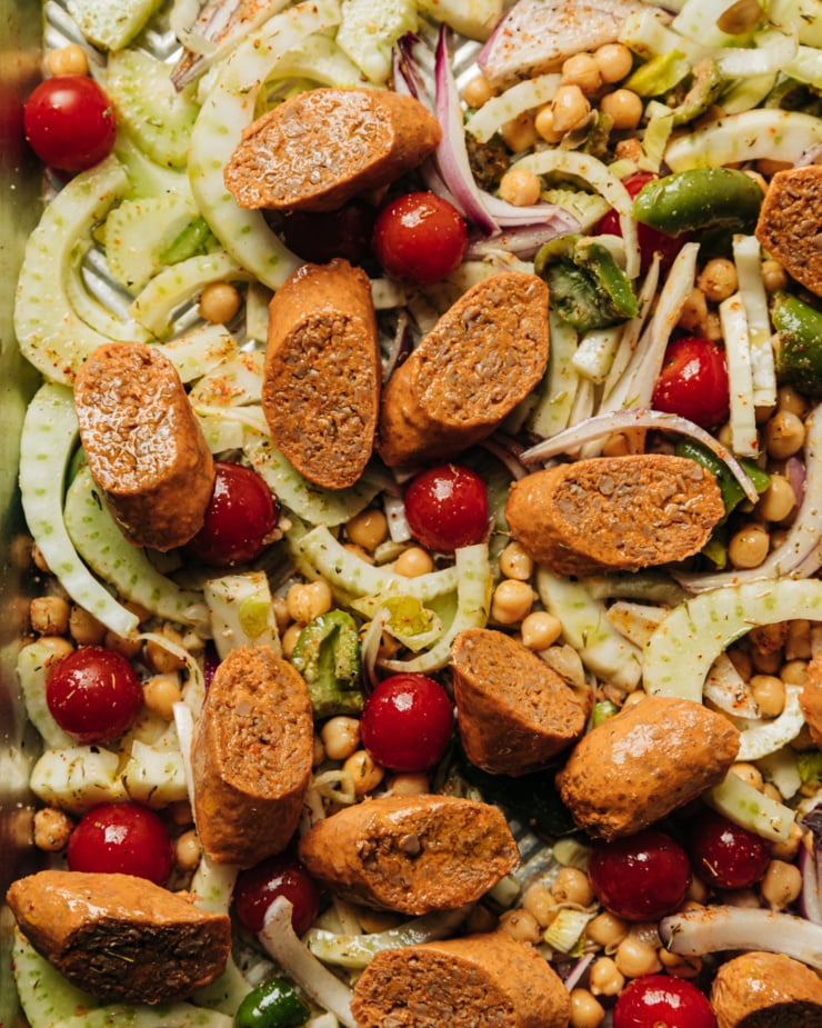 An overhead shot shows sliced vegan sausage on top of a mix of sliced fennel, cherry tomatoes, sliced red onion, chickpeas, and torn green olives.