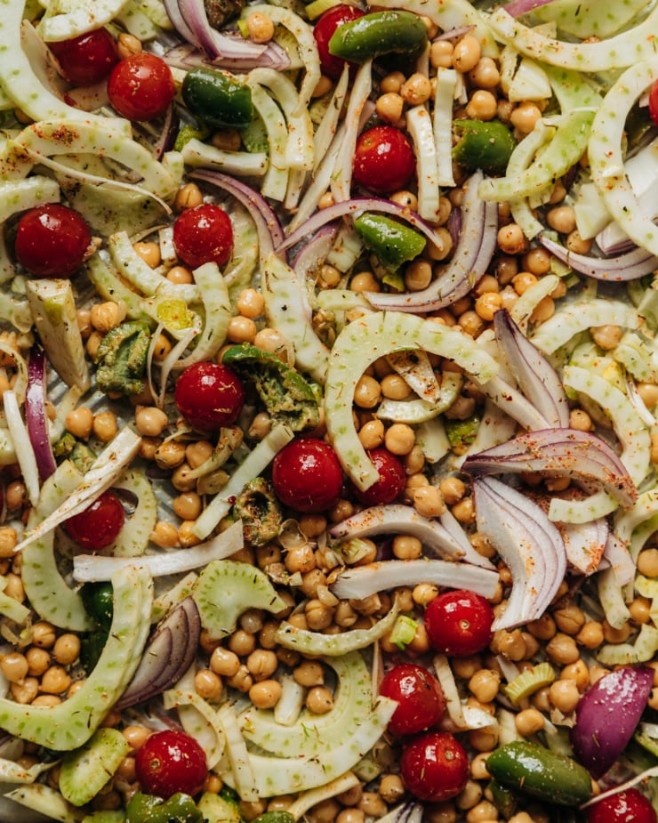 An up close, overhead shot of chickpeas, cherry tomatoes, sliced fennel, green olives, and sliced red onion on a baking sheet. Everything is dusted with spiced, salt, and pepper and thoroughly mixed up.